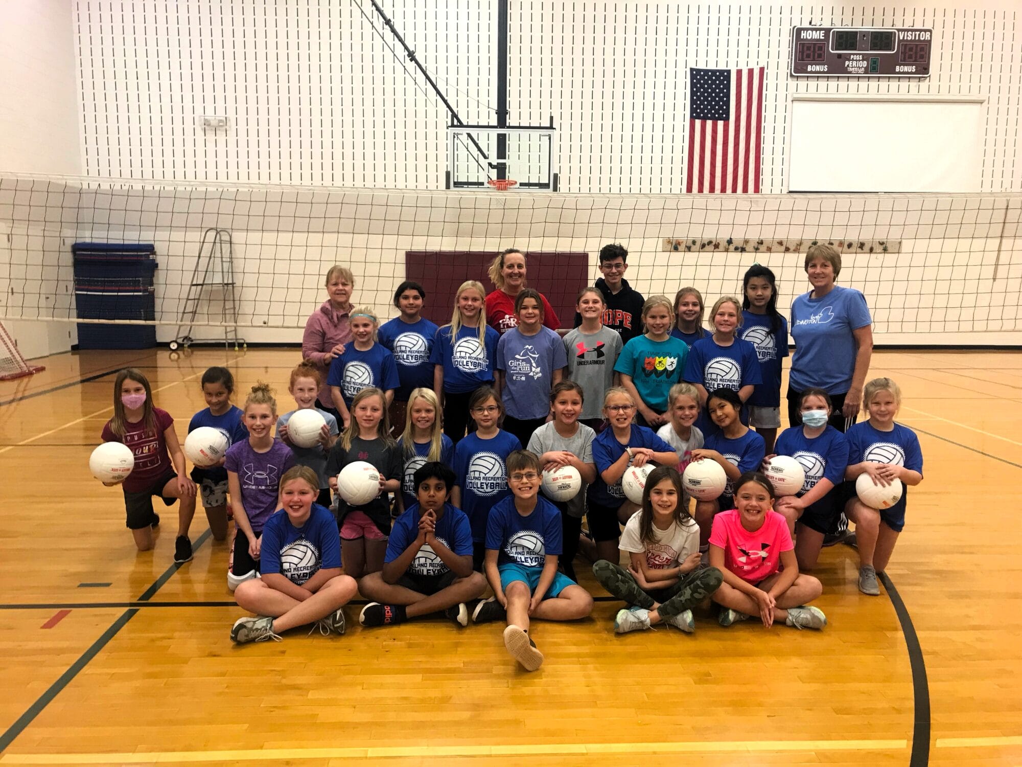 group of about 25 kids posing for a group picture with volleyballs in their hands in a gymnasium
