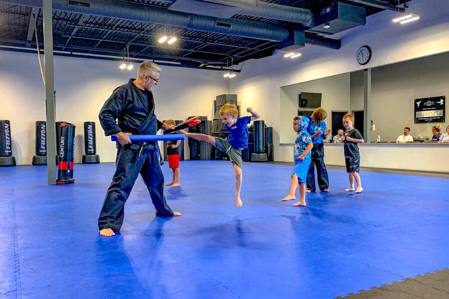 a martial arts instructor with a pool noodle in his hand, while a student kicks the noodle mid-air