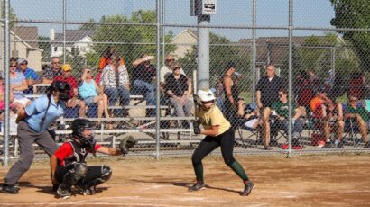 A teenage girl at bat during a softball game. The catcher is located to her left, as well as an official and a crow of people behind the fence.