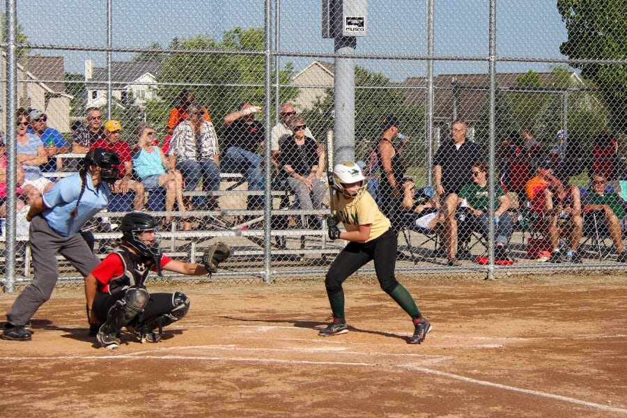 A teenage girl at bat during a softball game. The catcher is located to her left, as well as an official and a crow of people behind the fence.